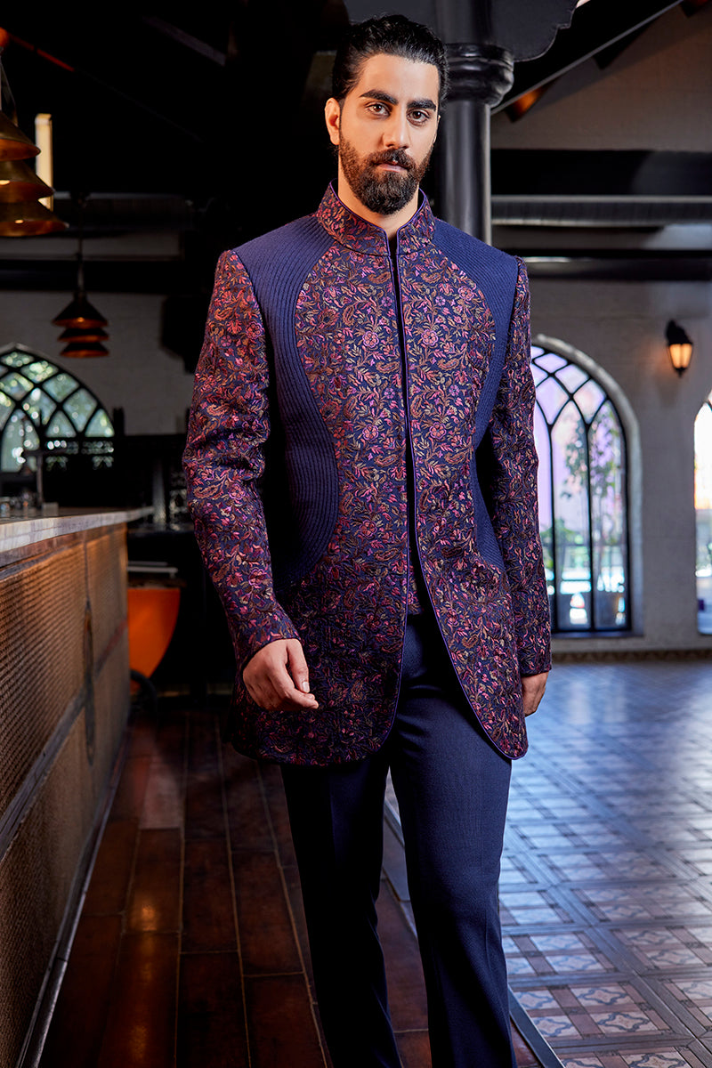 A man in a navy blue multi-coloured Kashmiri Indo-Western suit from BenzerWorld ethnicwear store poses in front of a counter.