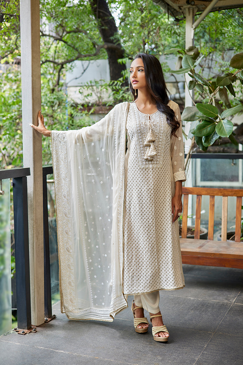 A woman poses in an off-white chanderi silk ladies suit from BenzerWorld ethnicwear store, as she leans against a column.