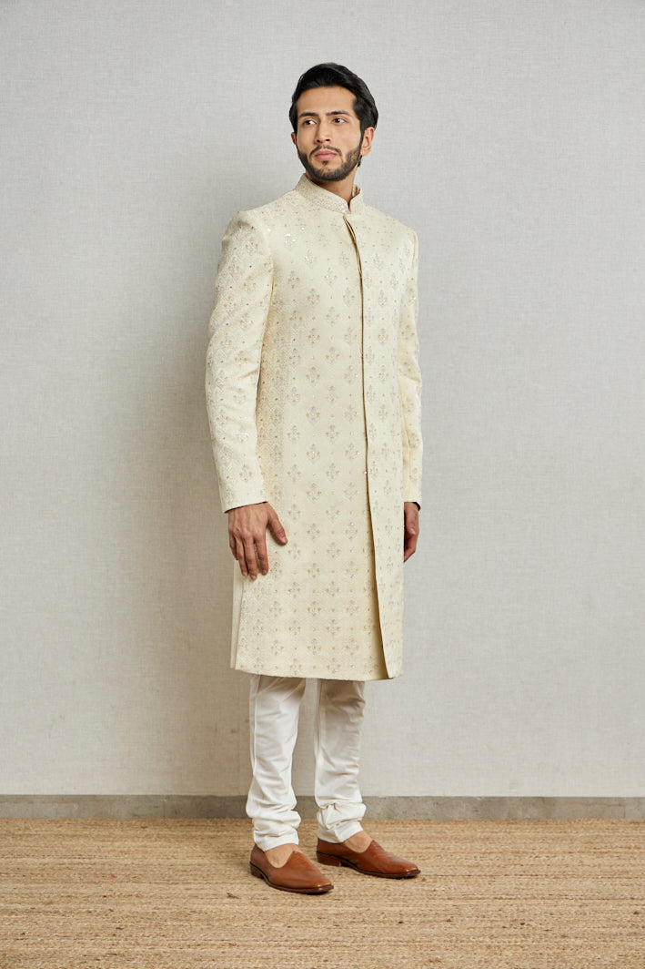 A man in an offwhite jacquard silk sherwani from BenzerWorld ethnicwear store poses in front of a wall.