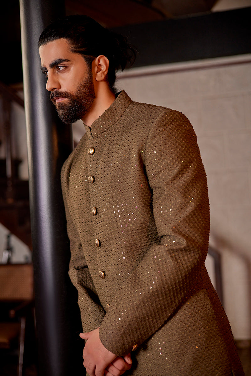 A man in an olive silk sherwani from BenzerWorld ethnicwear store poses in front of a pole.