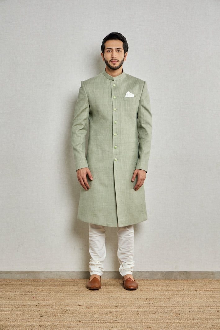 A man in a pista green silk sherwani from BenzerWorld ethnicwear store poses in front of a wall.