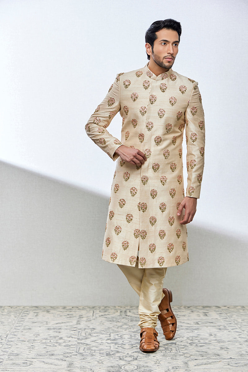A man poses in a powder peach raw silk sherwani from BenzerWorld ethnicwear store.