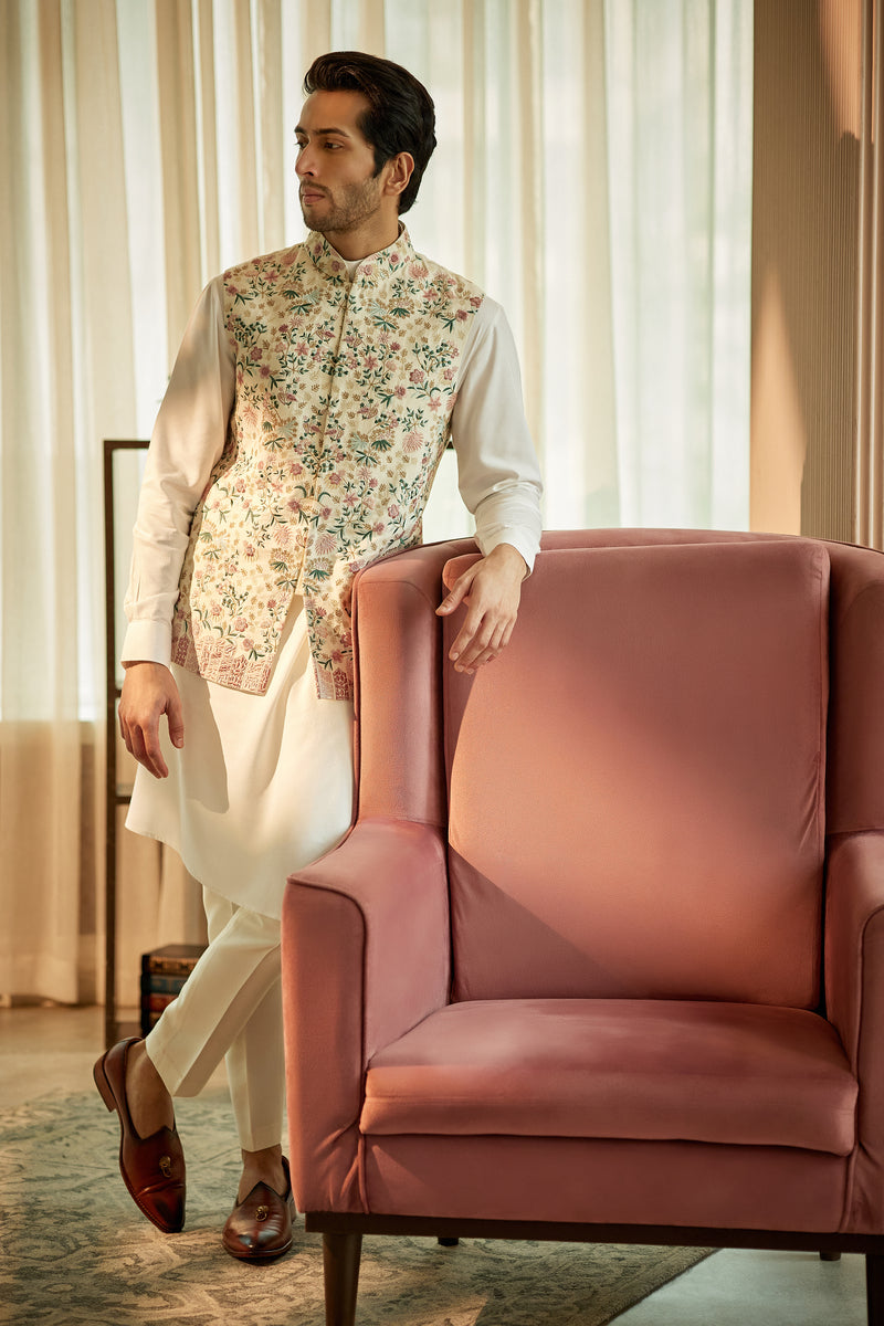 A man in a white matka silk kurta and bundi set from BenzerWorld ethnicwear store poses near a red chair.