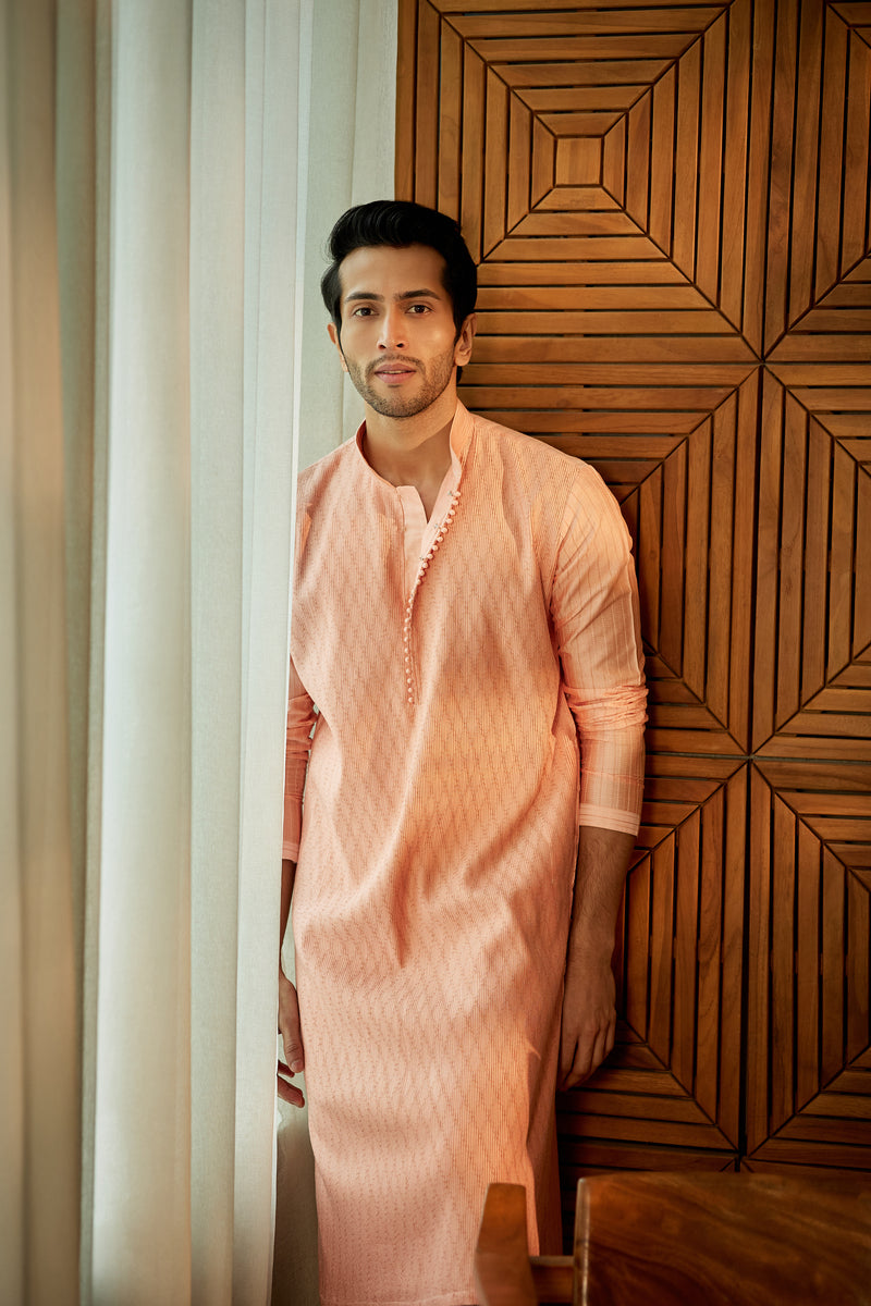 A man in a peach cotton silk kurta set from BenzerWorld ethnicwear store poses in front of a wood-panelled wall.