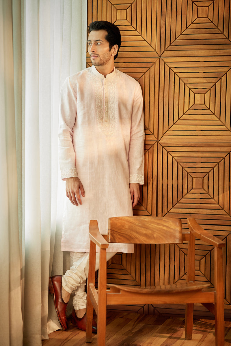 A man in a powder pink linen kurta set from BenzerWorld ethnicwear store poses in front of a wooden chair and wall.