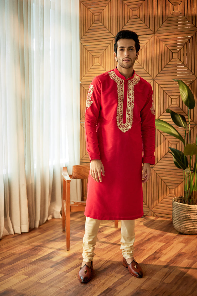 A man in a red cotton silk kurta set from BenzerWorld ethnicwear store poses in front of a wooden chair and wall.