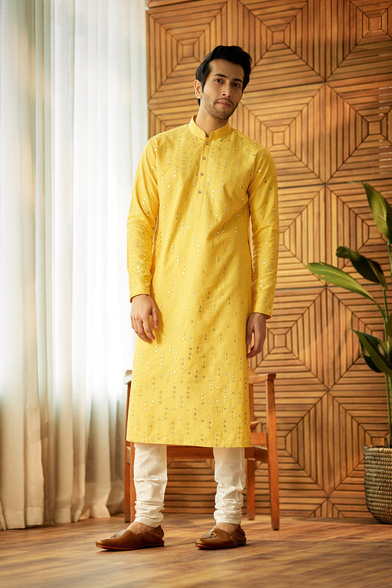 A man in a yellow chanderi silk kurta set from BenzerWorld ethnicwear store poses in front of a wooden chair and wall.