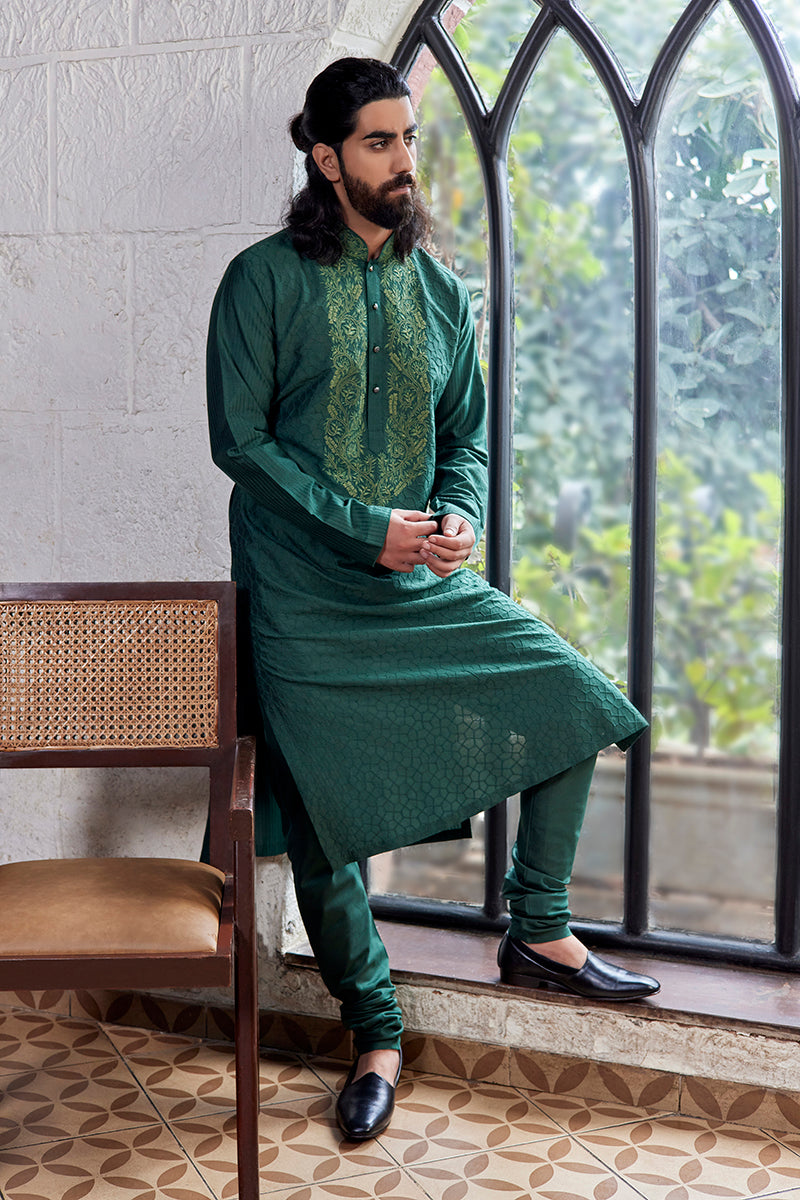 A man in a dark green silk kurta set from BenzerWorld ethnicwear store poses in front of an arched window.