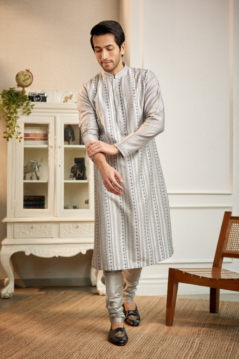 A man in a light grey chanderi silk kurta set from BenzerWorld ethnicwear store poses while adjusting his sleeve.