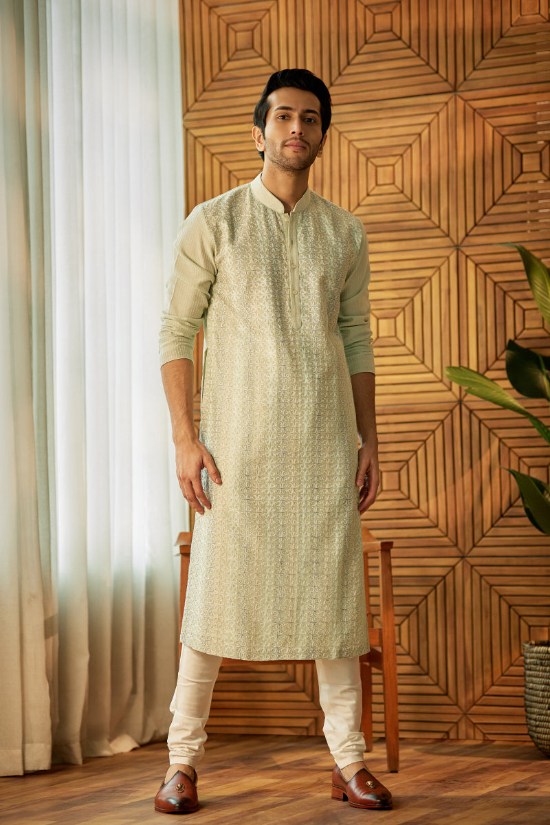 A man in a mint green chanderi silk kurta set from BenzerWorld ethnicwear store poses in front of a wooden chair.