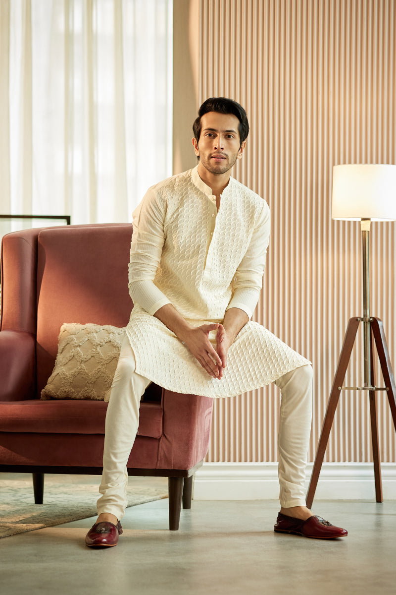 A man in a white cotton silk kurta set from BenzerWorld ethnicwear store poses on a red chair's armrest.